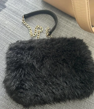 Svart fluffig kuvertväska Don Donna - Svart kuvertväska från Don Donna med mjuk, fluffig pälsimitation och guldfärgad kedja som handtag. Väskan har en snygg dragkedja i guld och detaljer i svart skinn. Perfekt storlek för mobilen och småsaker, med innerfack och logotyp på insidan.