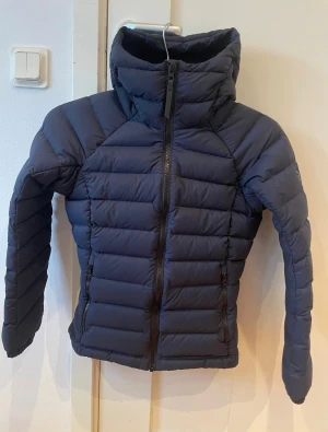 Mörkblå pufferjacka med huva - Snygg mörkblå pufferjacka med hög krage och stor huva i mycket fint skick! Jackan har horisontella quiltade sektioner, två sidofickor med dragkedja och en frontdragkedja. Perfekt för kalla dagar och har en clean, modern look som funkar till streetwear.
