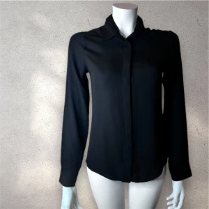 Svart långärmad skjorta - Stilren svart skjorta med klassisk krage och dolda knappar framtill. Skjortan har lång ärm och är tillverkad i ett tunt, lätt och något transparent polyestertyg. Perfekt för dig som gillar en clean och enkel look. #sjorta 