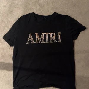 Amiri tröja  - Svart t-shirt från AMIRI med stort tryck över bröstet i beige och rosa toner. Klassisk rund hals och korta ärmar. Perfekt för dig som gillar streetwear och vill ha en statement piece i garderoben.