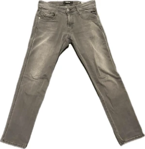 Replay Anbass grå jeans slim fit - Snygga grå Replay Anbass jeans med slim fit och klassisk femficksdesign. Jeansen har diskreta slitningar, raka ben och detaljerad brodering på bakfickorna. Perfekt för dig som gillar stilrena och moderna jeans med en cool touch.