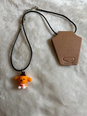 Halsband med Pompompurin-berlock - Svart halsband med en söt Pompompurin-berlock. Berlocken är formad som en liten hund med brunt hatt och röda detaljer. Perfekt accessoar för dig som gillar kawaii-stil och Sanrio-karaktärer.