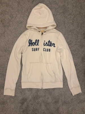 Vit Hollister hoodie - Vit hoodie från Hollister med blå broderad text 'Hollister Surf Club' på bröstet. Modellen har huva, två framfickor och dragkedja hela vägen. Tillverkad i mjuk bomullsblandning, perfekt för en chill och avslappnad stil. 