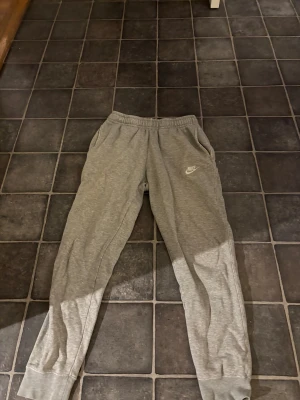 Grå mjukisbyxor från Nike, storlek S - Grå mjukisbyxor från Nike med klassisk logga på vänster ben. Byxorna har elastisk midja med snörning, sidofickor och en bakficka med lock. Tillverkade i mjukt bomullsmaterial för extra komfort. Perfekta för chill eller träning.