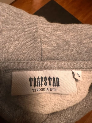Grå Trapstar hoodie med röd logga - Säljer en grå hoodie från Trapstar i storlek S. Tröjan har en stor röd och vit Trapstar-logga broderad på bröstet, snörning i huvan och ribbade muddar vid ärmar och nederkant. Materialet är mjukt och känns som bomull eller polyester.