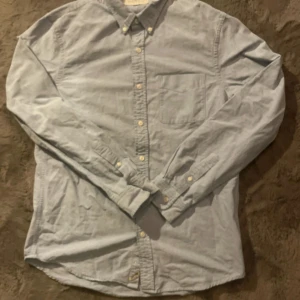 Ljusblå skjorta från Ralph Lauren - Snygg ljusblå skjorta från Denim & Supply Ralph Lauren med klassisk krage och bröstficka. Skjortan har långa ärmar, knappar framtill och en diskret logotyp vid nederkanten. Perfekt för dig som gillar stilrena och tidlösa plagg.