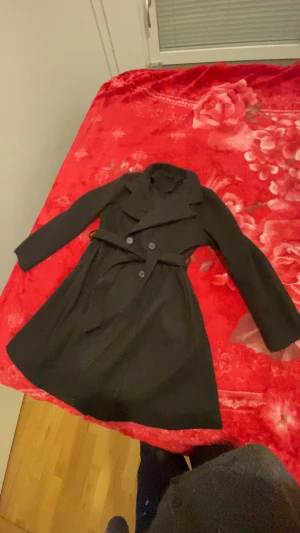 Svart kappa med bälte och knappar - Snygg svart kappa med klassisk krage och bälte i midjan. Dubbelknäppt framtill med stilrena knappar och långärmad design. Perfekt för dig som gillar en clean och elegant look under höst och vinter.