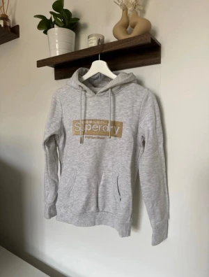 Ljusgrå Superdry hoodie med guldtryck - •Ljusgrå hoodie från Superdry med guldigt broderad logga på bröstet.  •I material av bomull och polyester.  ❗️Har en obehandlad fläck på ärmen, säljs därför billigare. I övrigt bra skick. 