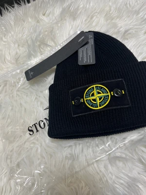 Svart ribbstickad mössa Stone Island - Svart ribbstickad mössa från Stone Island med klassisk logotyp-patch framtill i gult och grönt, fäst med två svarta knappar. Mössan är tillverkad i mjukt material och har en bred uppvikt kant för extra stilpoäng.