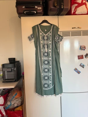 Grön långklänning med broderi - Säljer en grön långklänning med vita och gröna broderade mönster framtill och på ärmarna. Klänningen har v-ringning med snörning och tofsar, samt korta ärmar med fransdetaljer. Materialet är mjukt och känns som bomull. Perfekt för dig som gillar boho-vibe och unika detaljer. Storlek One Size men passar kanske en L/XL mer. Mycket fint skick, jag hittar inga synliga anmärkningar eller defekter. 
