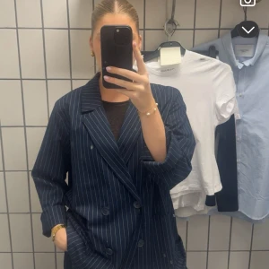 Mörkblå randig kavaj - Säljer en mörkblå kavaj med vita smala ränder och dubbelknäppning. Kavajen har breda slag, två framfickor och en klassisk, rak passform. Perfekt för dig som gillar en stilren och trendig look.