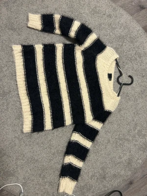Randig stickad tröja från KappAhl - Mysig stickad tröja från KappAhl i storlek M. Tröjan har breda svarta och beige ränder, rund hals och långa ärmar. Den grova stickningen ger en chill vibe och passar perfekt när du vill vara både bekväm och snygg.