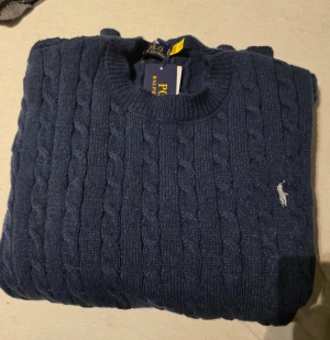 Stickad tröja  - Snygg marinblå stickad tröja  med klassiskt kabelstickat mönster och rund hals. Tröjan har en liten broderad logga i vitt på bröstet och ribbade muddar vid hals. ärmslut och nederkant. Perfekt för dig som gillar stilrena och tidlösa plagg.se mindre