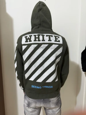 Off-White grön hoodie med tryck - Säljer en grön hoodie från Off-White med vit diagonaltryck på ryggen och ärmarna samt texten 'WHITE' och 'SEEING THINGS' på baksidan. Framsidan har Off-White-logga och en stor magficka. Hoodien har huva och ribbade muddar.