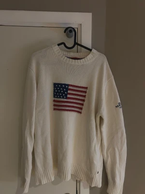 Vit stickad GANT tröja med USA-flagga - Priset kan diskuteras. Vit stickad tröja från GANT med broderad amerikansk flagga på bröstet och mörkblå text 'GANT NEW HAVEN 1949' på ärmen. Tröjan har rund hals, ribbade muddar och är långärmad. Materialet är 100% bomull, perfekt för dig som gillar klassisk college-stil.