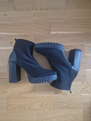 Svarta stövletter med klack från Zara - Säljer ett par svarta stövletter från Zara med chunky hög klack och grov plattformsula. Skorna har dragkedja framtill och är gjorda i syntetmaterial med detaljer i skinnimitation. Rund tå och cool urban vibe, perfekta för dig som gillar statement boots.