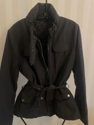 Svart parkajacka med bälte st.36 - Svart parkajacka från Ann Christine med hög krage, dragkedja och knappar framtill. Jackan har stora fickor med tryckknappar, justerbart bälte i midjan och långa ärmar. Materialet är polyester och jackan har en clean, stilren look som passar till det mesta.