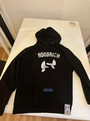 Svart hoodie från Hoodrich med tryck - Svart hoodie från Hoodrich med stor vit logga och två vita duvor på framsidan. Baksidan har ett stort tryck med fler duvor och blå texten 'From Nothing To Something'. Huva med snörning och känguruficka framtill. Mjuk bomullskänsla, perfekt för streetwear. Har knappt använt den bara legat hemma.