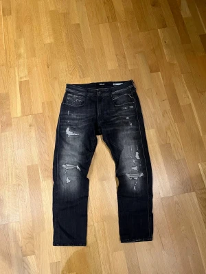 Replay Anbass svarta slim jeans 31 - Svarta Replay Anbass jeans i slim fit med slitna detaljer och snygg tvätt. Klassisk femficksmodell med dragkedja och knapp, samt Replay-logga på bakfickan. Jeansen har en modern look med distressed effekter och passar dig som gillar streetstyle.