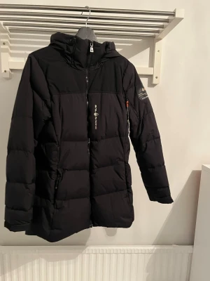 Svart dunjacka från Sail Racing - Svart lång dunjacka från Sail Racing med huva och dragkedja framtill. Jackan har diskreta loggor på bröstet och ärmen, samt praktiska fickor med dragkedja. Perfekt för kalla dagar med sin quiltade design och stilrena look.