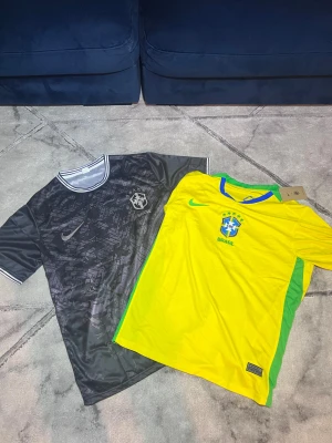 Brasilien Nike fotbollströjor - Två snygga Brasilien fotbollströjor från Nike. Ena är klassiskt gul med gröna detaljer och blå krage, den andra är svart med subtilt mönster och vita detaljer. Båda har Brasilien-emblem på bröstet och är gjorda i lätt polyester som andas.