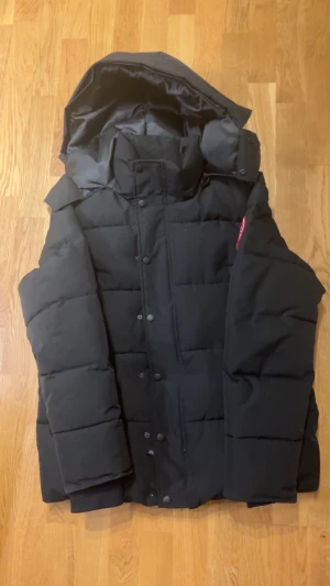 Svart dunjacka från Canada Goose S - Säljer en svart dunjacka från Canada Goose i storlek S/M. Jackan har en rejäl huva, klassisk patch på ärmen och stängs med både dragkedja och knappar. Fodret är glansigt och jackan är riktigt tjock och varm, perfekt för kalla vinterdagar. 