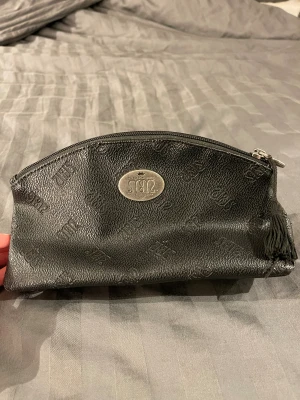 Svart plånbok från Maria Montazami - Svart necessär/plånbok från Maria Montazami 1812 Collection med präglad logga över hela ytan och en silverfärgad metallbricka framtill. Väskan har dragkedja och en liten tofs som detalj. Tillverkad i syntetmaterial med stilren look.