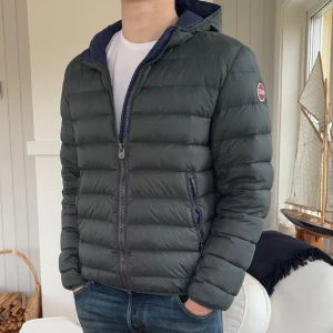 Mörkgrå Colmar pufferjacka med huva - Säljer en snygg mörkgrå pufferjacka från Colmar med huva och två sidofickor med dragkedja. Jackan har ett quiltat mönster och Colmar-logga på ärmen. Perfekt för kalla dagar och har en modern, clean look. Passar dig som gillar streetwear och funktion.