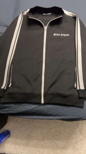 Palm Angels kofta - Svart track jacket från Palm Angels med vit dragkedja och vita ränder längs ärmarna. Jackan har hög krage och logga tryckt på bröstet. Tillverkad i mjuk polyester för en sportig och clean look.