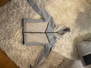 Nike tech flecce, Blå och vit  - Nike tech flecce i ljusblå och vit med svart dragkedja och detaljer. Jackan har huva, två stora fickor framtill och en sportig look med logga på bröstet. Perfekt för dig som gillar streetwear och vill ha en snygg jacka till vardags. Lite smutsig på framsidan.