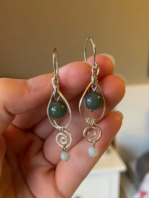 Handgjorda örhängen med grön sten - Unika örhängen i silverfärgad metall med spiralformad design och två gröna pärlor i varje örhänge. Den större pärlan sitter i mitten av en droppformad ram och en mindre ljusgrön pärla hänger längst ner. Perfekta för dig som gillar boho eller naturinspirerad stil.