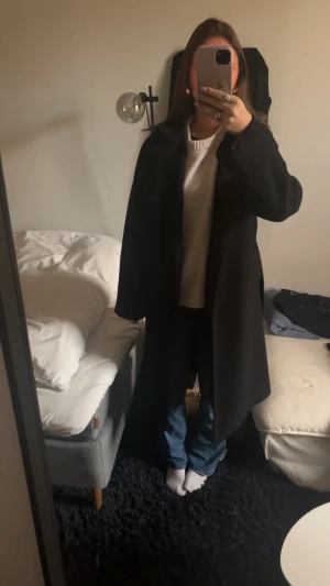 Svart kappa med bälte och krage - Säljer en svart kappa med bälte i midjan. Jackan har en klassisk, rak och lite oversized passform som ger en chill vibe. Materialet känns tjockt och varmt, perfekt för kyliga dagar. Enkel och stilren design som funkar till jeans och sneakers. 