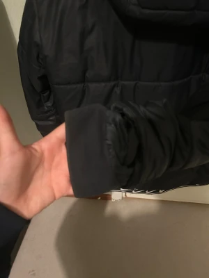 Svart pufferjacka från Nike M - Svart pufferjacka från Nike med huva och snörning. Jackan har dragkedja framtill, vita Nike-loggor på bröstet och insidan, samt coolt mönster med swoosh längs insidan. Perfekt för kalla dagar och har en modern, sportig vibe.