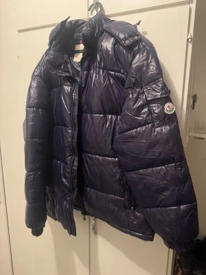 Moncler Maya Navy Blue (XL-XXL) - Säljer en mörkblå pufferjacka från Moncler med klassisk quiltad design och hög krage. Jackan har dragkedja framtill, två sidofickor med dragkedja och Moncler-logga på ärmen. Materialet är glansigt syntet. Passar någon som har XL-XXL, använd endast 1 gång då den var för stor för mig som har L-XL vanligtvis