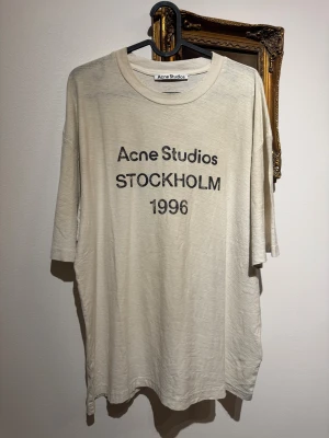Vit Acne Studios t-shirt storlek M 1996 Stockholm - Vit t-shirt från Acne Studios med tryck 'STOCKHOLM 1996' på bröstet. Modellen är kortärmad och har en klassisk rund hals. Materialet består av 85% bomull och 15% hampa, vilket ger en mjuk och lätt känsla. Perfekt för dig som gillar stilrena och trendiga plagg.