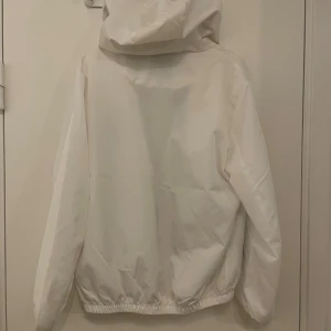 Vit vindjacka från Moncler med luva - Säljer en stilren vit vindjacka från Moncler med luva och svarta dragkedjor. Jackan har elastisk nederkant, två fickor med dragkedja och Moncler-logga på bröstet och ärmen. Perfekt för dig som vill ha en clean look med exklusiv känsla.