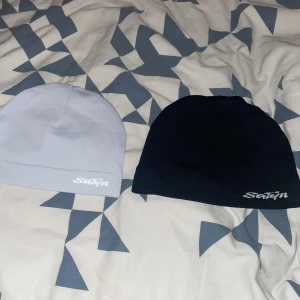 Satinmössa i svart och ljusgrå/vit 1 för 300kr - Två stilrena mössor från Satin, en i svart och en i ljusgrå. Utsidan är slät och enkel, medan insidan är fodrad med glansigt satinmaterial som är skonsamt mot håret. Perfekt för dig som vill ha en clean look och samtidigt ta hand om håret.