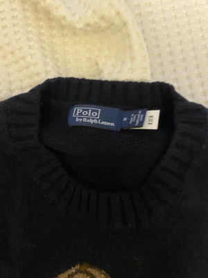 Stickad tröja med björn från Polo Ralph Lauren - Mörkblå stickad tröja från Polo Ralph Lauren med ikonisk björn på framsidan. Tröjan har rund halsringning och långa ärmar. Perfekt för dig som vill ha en klassisk och lekfull look med preppy-vibe.