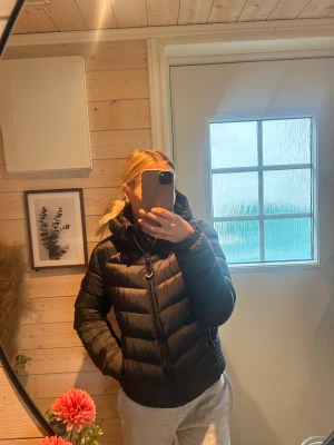 Svart pufferjacka Superdry  - Svart pufferjacka från Superdry Fuji Series med huva och dragkedja framtill. , två fickor med dragkedja och Superdry-logga på sidan, bara använd en gång