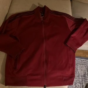 Vinröd Adidas ziptröja XL - Säljer en vinröd ziptröja från Adidas i storlek XL. Tröjan har klassiska svarta ränder längs ärmarna, ribbade muddar och hel dragkedja framtill. Perfekt för dig som gillar sportig stil och vill ha något bekvämt med snygg design.