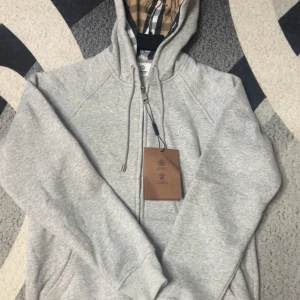 Grå hoodie från Burberry med dragkedja - Snygg grå hoodie från Burberry med klassiskt rutigt foder i huvan. Tröjan har dragkedja framtill, snörning vid huvan och ribbade muddar. Tillverkad i 100% bomull för skön komfort. Perfekt för dig som gillar stilrena och lyxiga detaljer.