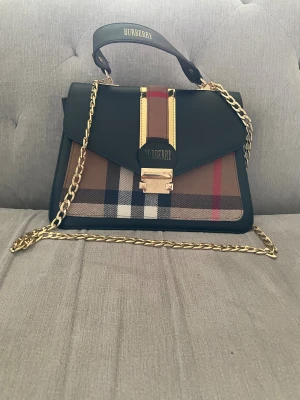 Rutig handväska från Burberry - Snygg handväska från Burberry med klassiskt rutigt mönster i brunt, svart, vitt och rött. Väskan har guldfärgade detaljer, kedja och ett elegant spänne framtill. Utsidan är i skinn och tyg, insidan är vinröd. Perfekt för dig som vill ha en ikonisk accessoar.
