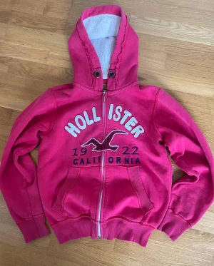 Rosa hoodie från Hollister med dragkedja - Snygg rosa hoodie från Hollister med broderad logga och text på bröstet. Tröjan har huva med vit foder, ribbade muddar och fickor framtill. Se bilderna för defekter!! ❤️❤️Storlek S men sitter ganska spänt så sku säga att de är mer som XS