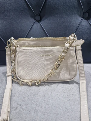 Beige axelväska från Valentino - Snygg beige axelväska från Valentino med lyxig guldfärgad kedja och präglad struktur som liknar reptil. Väskan har två fack med dragkedja och detaljer i guld, perfekt för att lyfta din outfit. Stilren design med Valentino-logga framtill.