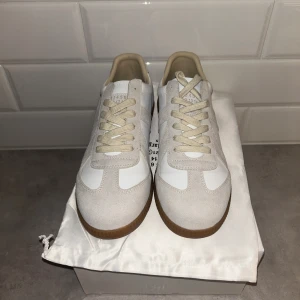 Maison Margiela sneakers beige/vit - Maison Margiela sneakers i beige och vitt med klassisk gummisula. Ovandel i skinn och mocka, neutrala färger och snygg retrostil. Perfekta för dig som gillar minimalistisk och stilren look. Snörning framtill och rund tå.