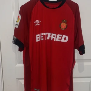 Röd Mallorca fotbollströja Umbro - Mallorca fotbollströja från Umbro i röd färg med svarta detaljer vid krage och ärmslut. Kortärmad modell med klubbmärke och sponsortryck på bröstet. La Liga-märke på ärmen. Tillverkad i lätt och ventilerande polyester, perfekt för match eller träning.