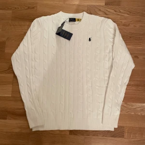 Vit kabelstickad tröja Polo Ralph Lauren - Snygg vit kabelstickad tröja från Polo Ralph Lauren med klassisk rund hals och broderad logga på bröstet. Tröjan har ribbade muddar vid ärmslut och nederkant, vilket ger en schysst passform. Perfekt för dig som gillar stilrena och tidlösa plagg.