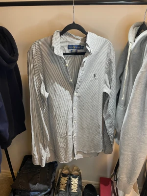 Randig skjorta från Ralph Lauren - Slim fit skjorta från Ralph Lauren i stretchig oxford-bomull. Skjortan är vit med smala svarta ränder, har klassisk krage, knappar framtill och broderad logga på bröstet. Långärmad och perfekt för dig som gillar stilren och clean look.