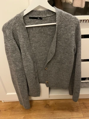 Grå stickad kofta från 157 - Mysig grå stickad kofta från 157 med tre knappar framtill. Koftan har lång ärm och en enkel, klassisk design som är lätt att matcha med olika outfits. Perfekt för kyliga dagar när du vill vara både bekväm och snygg. Stolek S