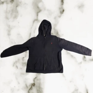 Svart hoodie från Polo Ralph Lauren  - Svart hoodie från Polo Ralph Lauren. Tröjan har huva, dragkedja framtill och två fickor. Materialet är mjuk bomull med insida i fleece. Klassisk röd Polo-logga på bröstet och långärmad passform. Perfekt för chill dagar och streetwear.